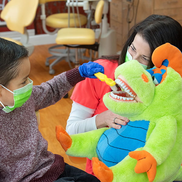A little boy wearing a green mask while brushing the teeth of a stuffed animal 