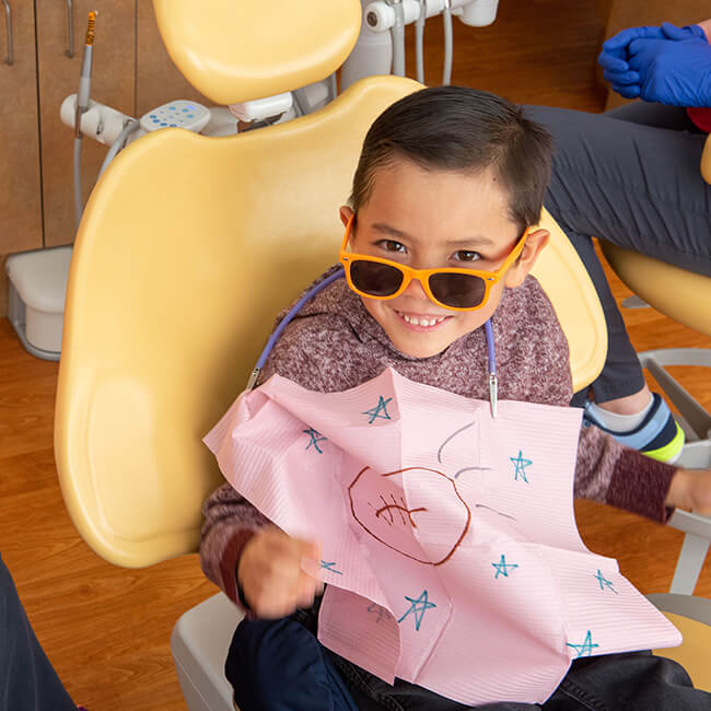 A little boy with white teeth sitting in a dental chair wearing a dental bib and a sunglass