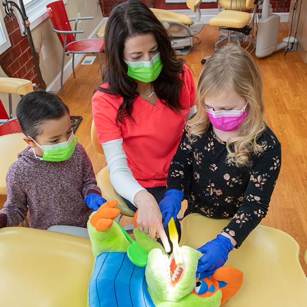 A team member teaching a little girl and a little boy how to brush teeth using a stuffed animal model lying in a dental chair