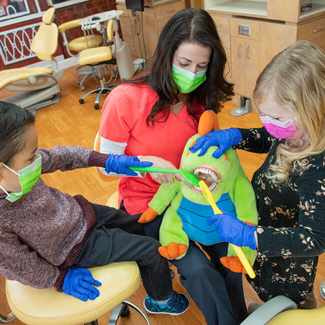A little boy and girl wearing a mask brushing the teeth of a stuffed animal using a toothbrush while a Doctor Karen is holding it
