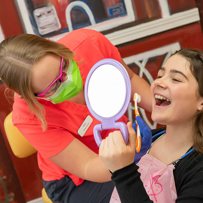 A little girl lying on a dental chair with mouth open while holding a purple mirror and a team member inserting a toothbrush into her mouth